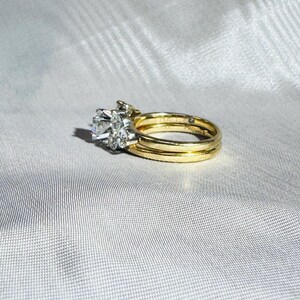 May include: A gold wedding ring set with a large, clear, round-cut diamond center stone and smaller accent diamonds. The set includes a matching band and a second, thinner gold band. The rings are displayed on a white, textured surface.