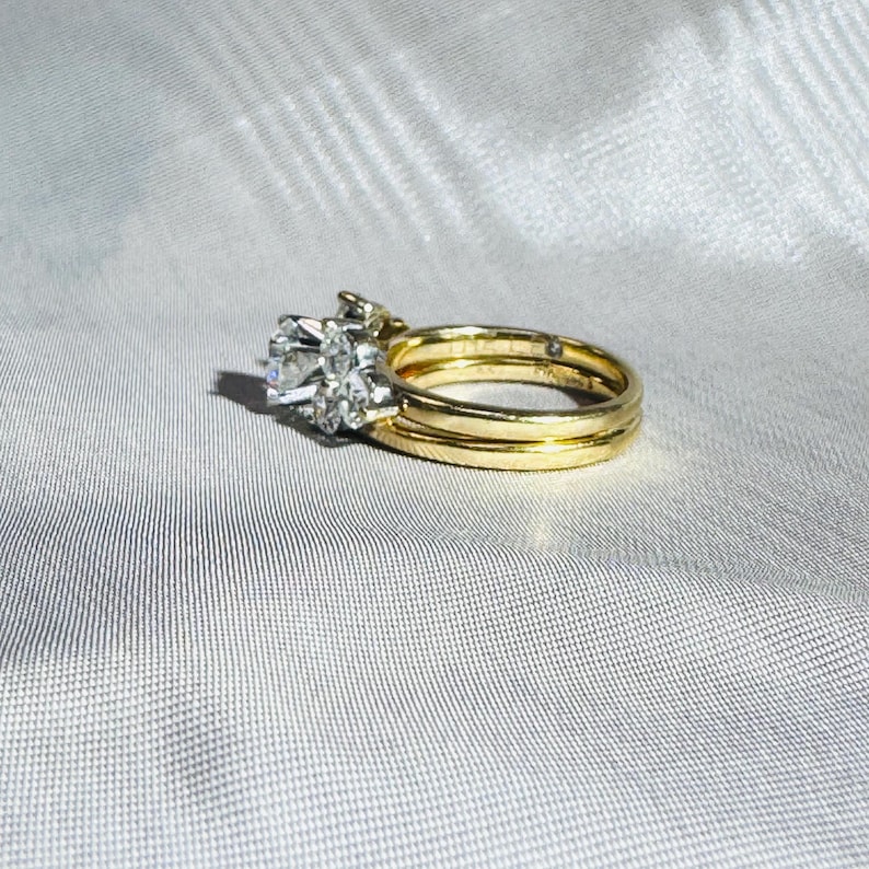 May include: A gold wedding ring set with a large, clear, round-cut diamond center stone and smaller accent diamonds. The set includes a matching band and a second, thinner gold band. The rings are displayed on a white, textured surface.
