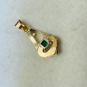 May include: A gold pendant with a small, square emerald gemstone. The pendant has a unique, openwork design with a loop at the top for a chain. The pendant is likely made of 18K gold.