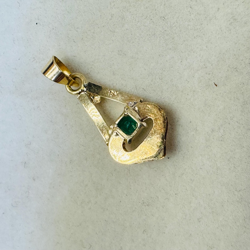 May include: A gold pendant with a small, square emerald gemstone. The pendant has a unique, openwork design with a loop at the top for a chain. The pendant is likely made of 18K gold.