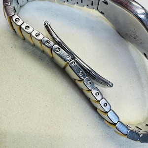 Pode incluir: Close-up de uma pulseira de rel&oacute;gio prateada e dourada. A pulseira apresenta uma s&eacute;rie de elos retangulares com bordas douradas e centros prateados. O fecho &eacute; prateado e curvo. A pulseira do rel&oacute;gio est&aacute; sobre uma superf&iacute;cie clara.