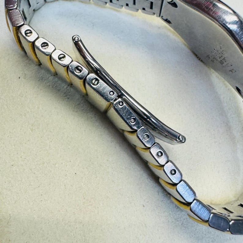 Pode incluir: Close-up de uma pulseira de rel&oacute;gio prateada e dourada. A pulseira apresenta uma s&eacute;rie de elos retangulares com bordas douradas e centros prateados. O fecho &eacute; prateado e curvo. A pulseira do rel&oacute;gio est&aacute; sobre uma superf&iacute;cie clara.