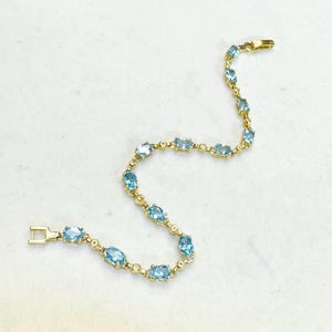 May include: A gold-toned bracelet featuring oval-shaped, light blue gemstones. The bracelet has a delicate design with a clasp closure. The gemstones are evenly spaced along the length of the bracelet, creating a sparkling, elegant look.