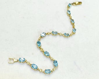 May include: A gold-toned bracelet featuring oval-shaped, light blue gemstones. The bracelet has a delicate design with a clasp closure. The gemstones are evenly spaced along the length of the bracelet, creating a sparkling, elegant look.