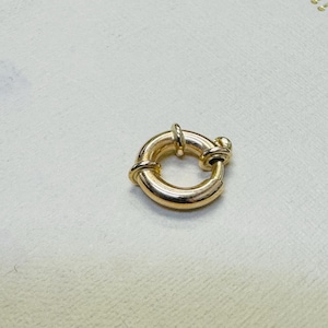 Puede incluir: Un cierre de resorte dorado pulido con un diseño circular y un pequeño mecanismo de palanca. El componente de joyería tiene una superficie metálica lisa y reflectante, con una abertura funcional para sujetar cadenas o colgantes.