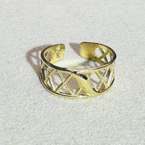 Puede incluir: Un anillo abierto de tono dorado que presenta un patrón repetitivo de celosía o forma de diamante en toda la banda. La superficie metálica tiene un acabado pulido y el anillo está diseñado con una abertura en la parte posterior para ajustar el tamaño.