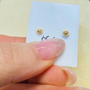 Op de afbeelding: Een paar kleine, goudkleurige oorstekers. Elke oorbel heeft een centraal verhoogd ontwerp omgeven door een kralenrand. De oorbellen worden gepresenteerd op een lichtblauwe kaart.