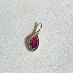 May include: A gold pendant with a marquise-cut, deep red gemstone. The pendant has a small loop at the top for a chain. The gemstone is set in a gold bezel, creating a striking contrast. The pendant is likely a piece of jewelry.