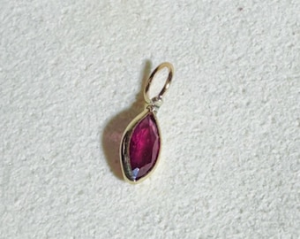May include: A gold pendant with a marquise-cut, deep red gemstone. The pendant has a small loop at the top for a chain. The gemstone is set in a gold bezel, creating a striking contrast. The pendant is likely a piece of jewelry.