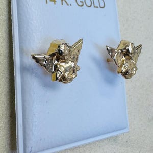 Op de afbeelding: Een paar goudkleurige engel oorknopjes. Elke oorbel heeft een gedetailleerd cherubijnontwerp met vleugels. De oorbellen worden gepresenteerd op een witte kaart, met de tekst "14K GOLD" bovenaan.