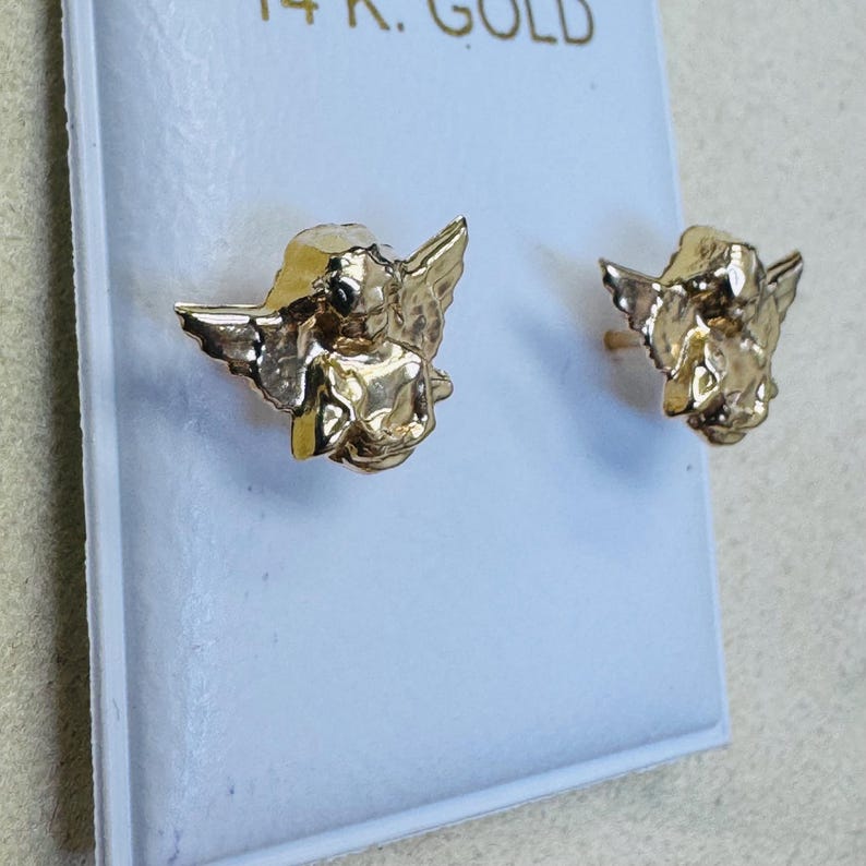Op de afbeelding: Een paar goudkleurige engel oorknopjes. Elke oorbel heeft een gedetailleerd cherubijnontwerp met vleugels. De oorbellen worden gepresenteerd op een witte kaart, met de tekst "14K GOLD" bovenaan.