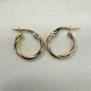 Puede incluir: Un par de pendientes de aro dorados. Cada pendiente tiene un diseño de cuerda trenzada y un cierre de bisagra. Los pendientes están hechos de un metal dorado brillante. Los pendientes se muestran sobre un fondo neutro.