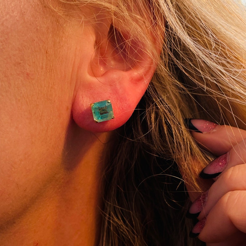 May include: A close-up of a woman's ear with a square emerald green gemstone earring. The earring is set in gold.