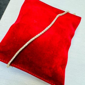 May include: A gold tennis bracelet with numerous small, square-cut, clear gemstones. The bracelet is displayed on a red velvet pillow, creating a contrast between the jewelry and the background. The bracelet is a delicate piece of jewelry.