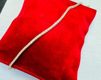 May include: A gold tennis bracelet with numerous small, square-cut, clear gemstones. The bracelet is displayed on a red velvet pillow, creating a contrast between the jewelry and the background. The bracelet is a delicate piece of jewelry.