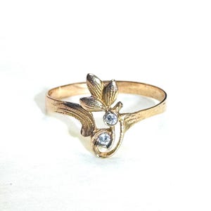 May include: A gold ring featuring a textured leaf-shaped design and two small, clear, round-cut stones set vertically. The band has a polished finish and tapers toward the decorative element, which is positioned at the center of the ring's top.