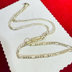 May include: A gold-colored Figaro chain necklace. The necklace is displayed on a white surface with a red background. The chain has a pattern of alternating elongated and round links. The necklace is a jewelry item.