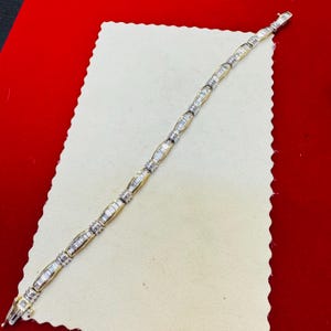 May include: A gold and silver bracelet with a series of rectangular and oval links. The bracelet is laid out on a white surface against a red background. The links are alternating in design.