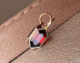 May include: A gold-colored pendant with a rectangular, faceted, deep red gemstone. The gemstone is set in a gold frame with a small loop at the top for a chain. The pendant is displayed on a textured, brown surface.