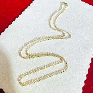 May include: A delicate gold-colored necklace with a fine chain composed of small, evenly spaced beads. The necklace is displayed on a white surface with a red border, creating a contrast that highlights the jewelry's details.