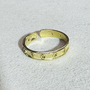 Puede incluir: Un anillo de metal en tono dorado con un diseño de banda abierta. La superficie exterior del anillo está decorada con un patrón repetitivo de estrías verticales intercaladas con pequeños recortes circulares que rodean toda la banda.