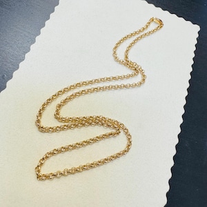 May include: A gold-colored chain necklace with a round link design. The necklace is displayed on a white surface with a scalloped edge. The chain is a warm, bright gold color and appears to be made of metal.
