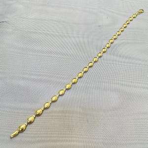 May include: A gold-toned chain bracelet featuring a series of repeating, smooth, marquise-shaped links connected by small circular rings. The bracelet is arranged in a straight line, showcasing a polished metallic finish and a uniform design.
