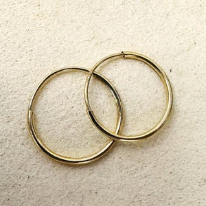 Puede incluir: Un par de pendientes de aro dorados. Los pendientes son circulares, con una superficie lisa y pulida. Los aros están entrelazados y el diseño general es simple y elegante. Los pendientes están sobre un fondo neutro.