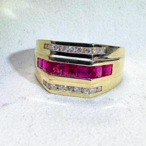 Op de afbeelding: Een gouden ring met een rij felrode edelstenen, waarschijnlijk robijnen, tussen twee rijen kleinere, heldere edelstenen. De ring heeft een rechthoekig ontwerp met een gepolijste afwerking, wat een klassieke stijl laat zien.