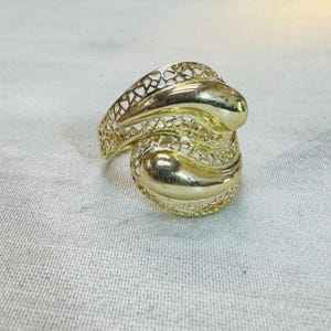 May include: A gold-colored ring with an openwork design. The ring features two rounded, smooth sections connected by a decorative, open filigree pattern. The ring is set against a neutral background.