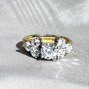 May include: A gold ring with a cluster of diamonds. The ring features a large, round diamond in the center, surrounded by smaller diamonds. The diamonds sparkle against the gold band, creating a luxurious look. The ring is set on a gray, textured surface.