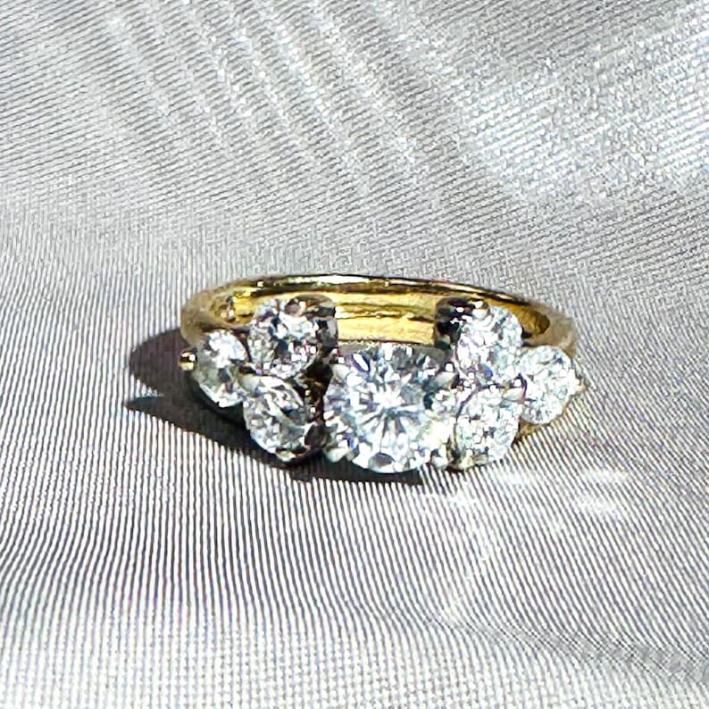 May include: A gold ring with a cluster of diamonds. The ring features a large, round diamond in the center, surrounded by smaller diamonds. The diamonds sparkle against the gold band, creating a luxurious look. The ring is set on a gray, textured surface.