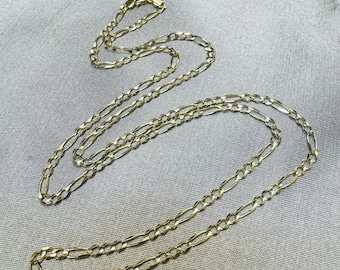May include: A long gold-toned figaro chain necklace featuring a repeating pattern of three small circular links followed by one elongated oval link. The metallic chain is arranged in a loose, overlapping loop formation against a neutral background.