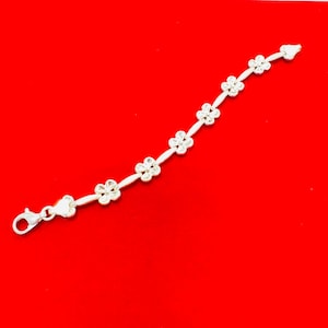 May include: A silver bracelet with a floral design. The bracelet features small, detailed flower charms connected by delicate links. The clasp is a lobster claw style. The bracelet is displayed against a bright red background.