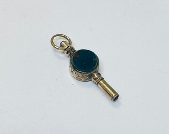 May include: A gold-toned watch key with a black and red stone set into the top. The key has a decorative floral design.