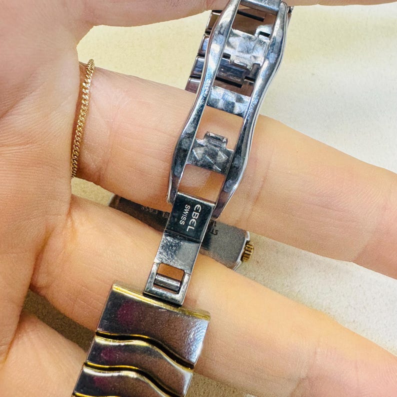 Pode incluir: Close-up de um fecho de rel&oacute;gio Ebel Swiss prateado e dourado. O fecho &eacute; retangular com o nome da marca "EBEL SWISS" gravado. A pulseira do rel&oacute;gio &eacute; feita de metal prateado. Uma corrente dourada &eacute; vis&iacute;vel no fundo.