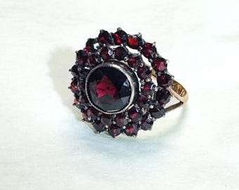 Puede incluir: Un anillo de oro con una gran piedra preciosa redonda de color rojo oscuro facetada en el centro, rodeada por dos círculos concéntricos de piedras preciosas más pequeñas, redondas y facetadas de color rojo oscuro en un diseño de racimo.