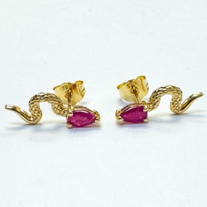 Può includere: Un paio di orecchini a lobo a forma di serpente color oro. Ogni orecchino presenta un corpo di serpente strutturato e una gemma rosa brillante a forma di goccia. Gli orecchini sono su uno sfondo bianco.