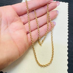 May include: A gold-colored chain necklace with a rounded link design. The necklace is held in a hand, showcasing its length and the details of the links. The chain is displayed against a neutral background.