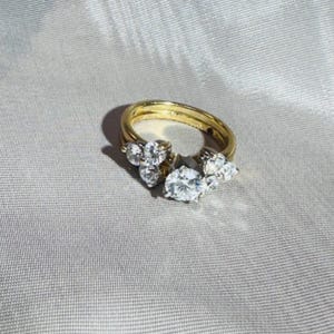 May include: A gold ring set with multiple clear, sparkling gemstones. The ring features a cluster of stones, including a large oval-cut center stone and smaller accent stones. The ring is set against a soft, neutral background.