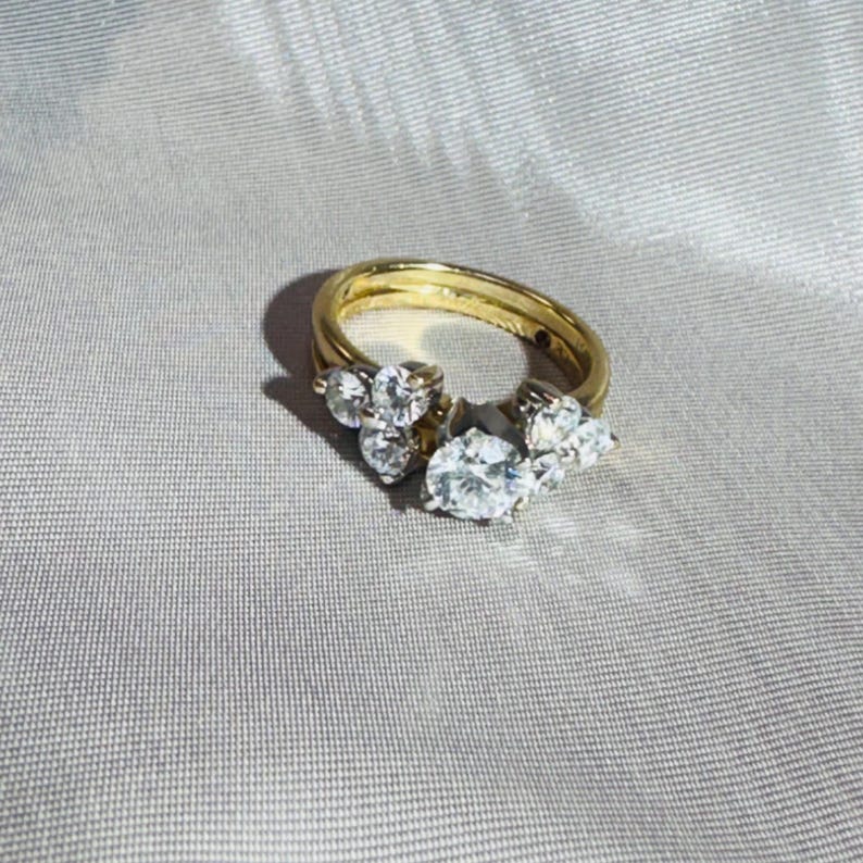 May include: A gold ring set with multiple clear, sparkling gemstones. The ring features a cluster of stones, including a large oval-cut center stone and smaller accent stones. The ring is set against a soft, neutral background.