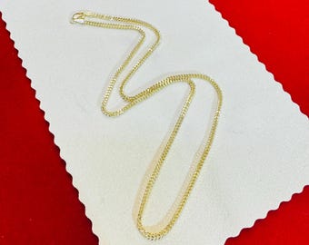 May include: A gold-colored chain necklace with a delicate, textured design. The necklace is displayed on a white, textured surface against a red background. The chain is likely made of gold or a gold-toned metal.