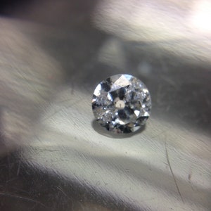 May include: A clear, round, faceted gemstone with a silver-toned setting. The gemstone is on a silver-colored surface.