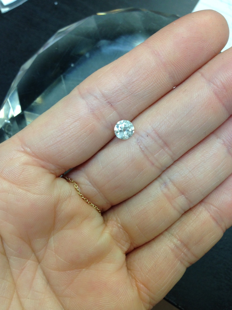 May include: A clear round gemstone with facets, held in a hand.