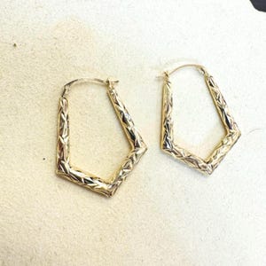Könnte beinhalten: Ein Paar goldfarbene, geometrische Creolen. Die Ohrringe haben ein strukturiertes, rautenförmiges Muster auf der Oberfläche. Die Form ist ein modifiziertes Rechteck mit abgewinkelten Seiten und einem Klappverschluss.