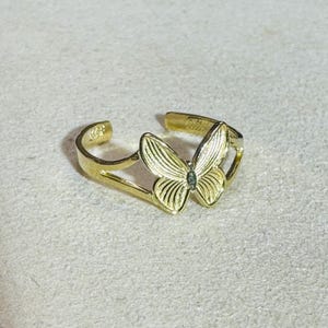 Puede incluir: Un anillo abierto de tono dorado que presenta un motivo central de mariposa con texturas detalladas en las alas. La banda es ajustable, con una abertura en la parte posterior, y la superficie metálica tiene un acabado pulido y brillante.