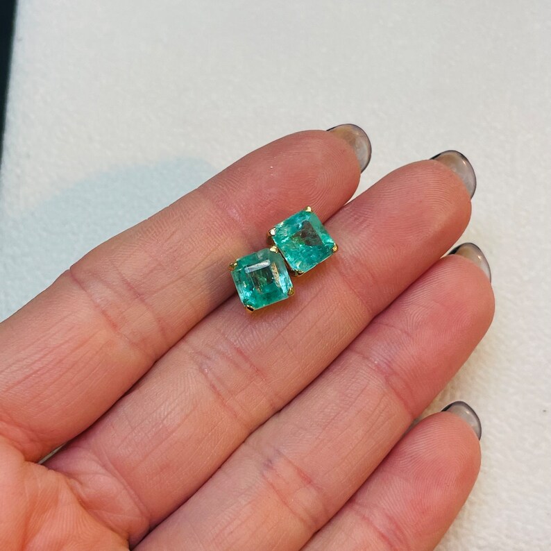 May include: A pair of emerald green gemstone earrings with gold settings. The earrings are square shaped and have a polished finish.