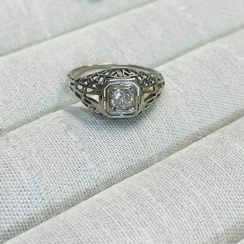May include: A delicate filigree ring with a square-shaped diamond set in a bezel setting. The ring is made of white metal and has a vintage style.