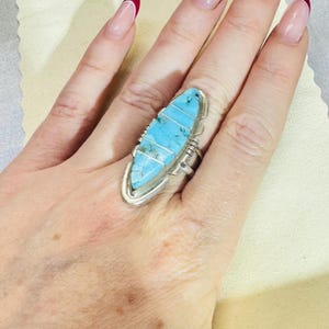 May include: A silver ring featuring a long, marquise-shaped inlay of light blue turquoise stone segments separated by thin silver bars. The metal setting has a polished finish with decorative notched edges along the sides of the stone inlay.