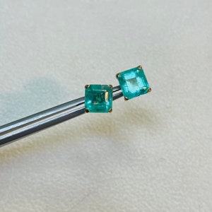 May include: A pair of emerald green square-shaped earrings with gold settings. The earrings are held by a pair of tweezers.
